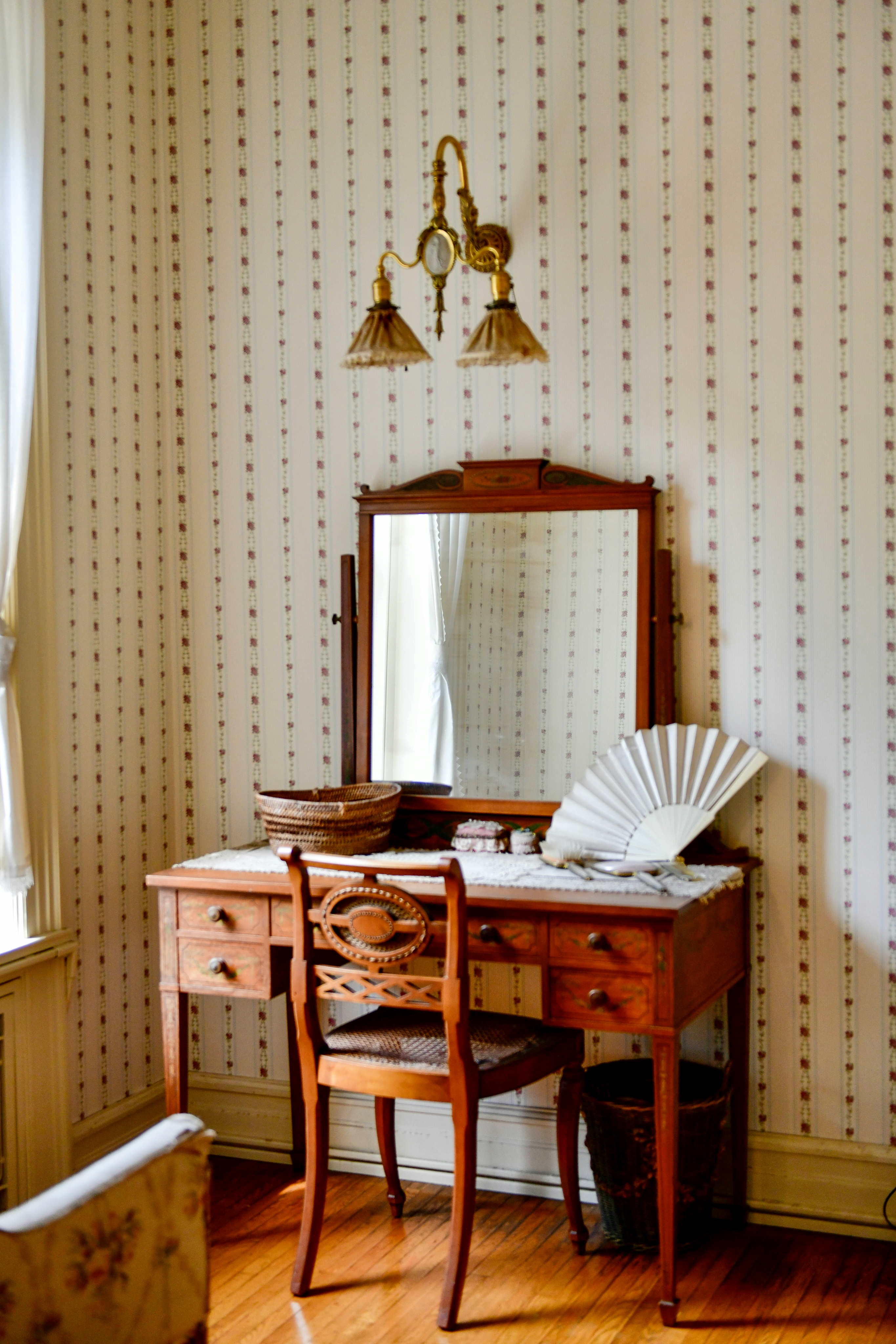 A vintage wooden desk with a mirror in the corner of a room with patterned wallpaper.